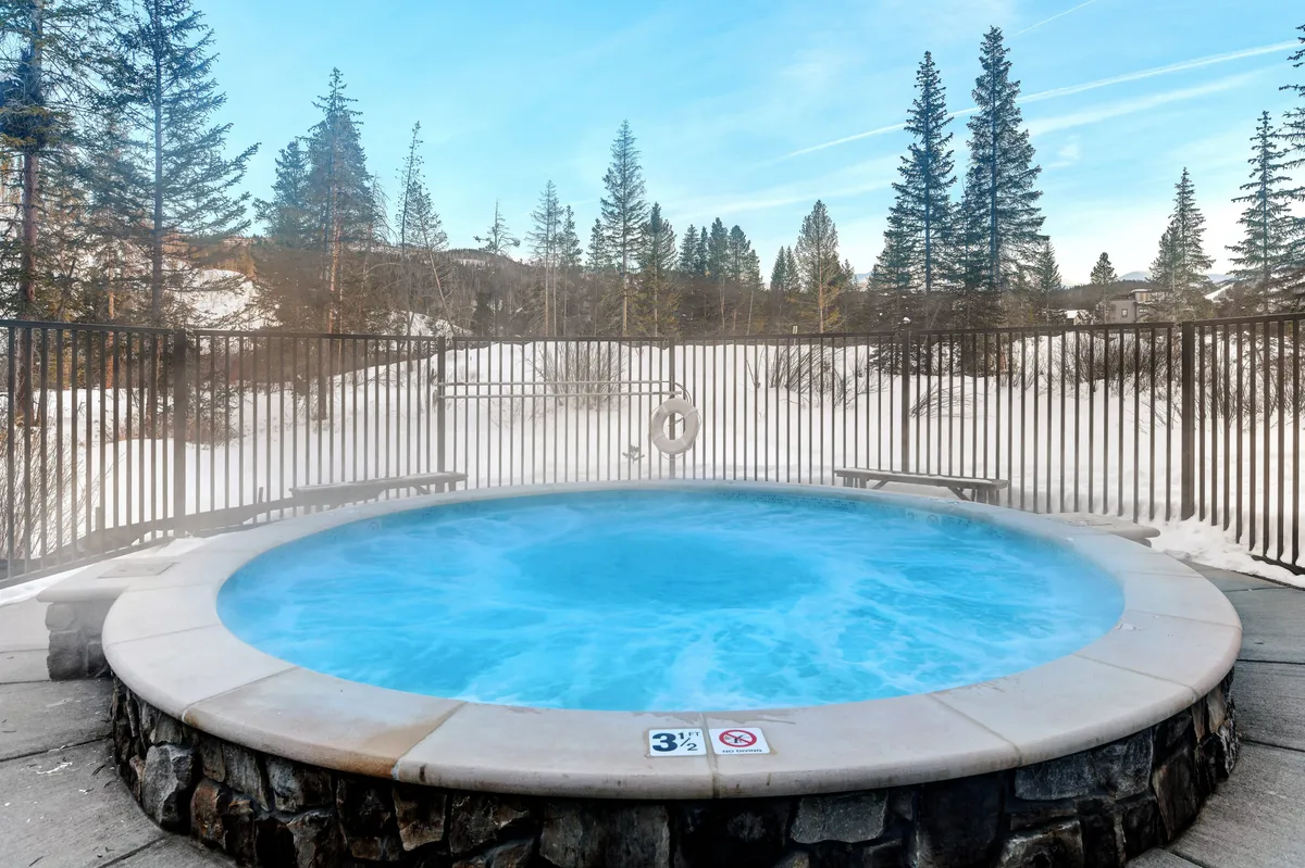 This exterior image features a hot tub surrounded by a snowy landscape and pine trees, with people relaxing and socializing. The serene setting reflects the perfect après-ski retreat, inviting guests to unwind after outdoor adventures.
