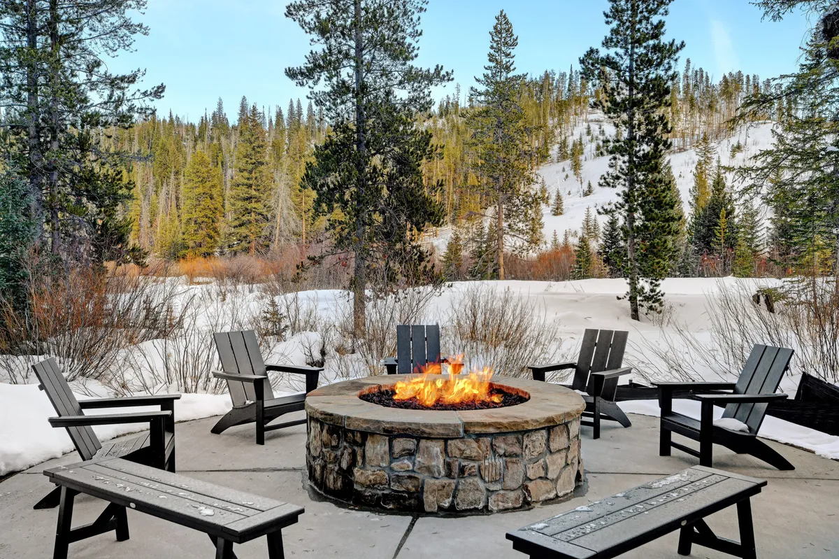 An outdoor fire pit glows warmly amid snow-covered ground and surrounding trees, where groups can gather and share stories or hot drinks against the alpine backdrop.