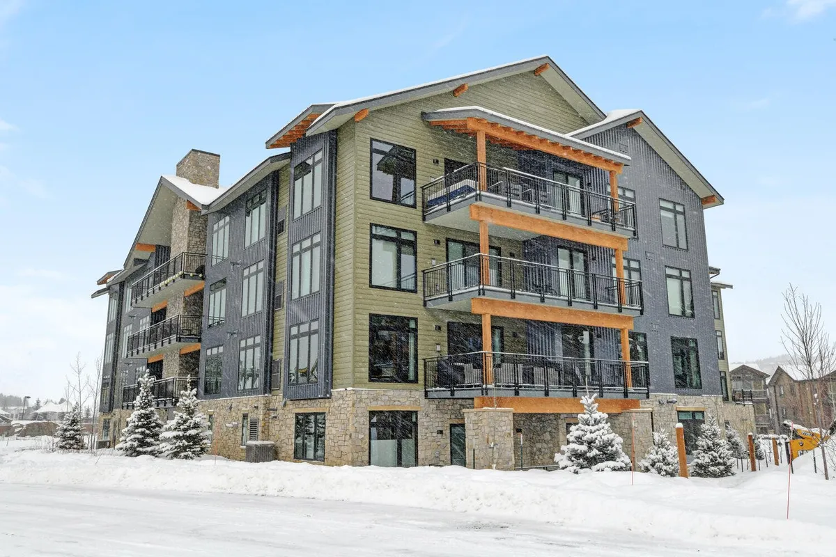 A striking exterior shot featuring the modern condo building with balconies overlooking a pristine, snow-covered landscape. The stone base of the building and warm lighting offer a welcoming feel, while the balconies hint at private outdoor spaces perfect for enjoying mountain views.