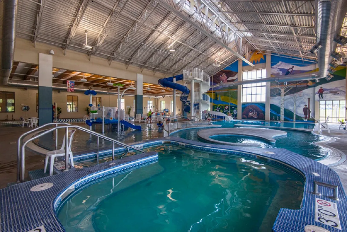 A vibrant indoor recreation center scene, featuring swimming pools, colorful water slides, playful murals, and lounge chairs filled with families and children enjoying themselves.