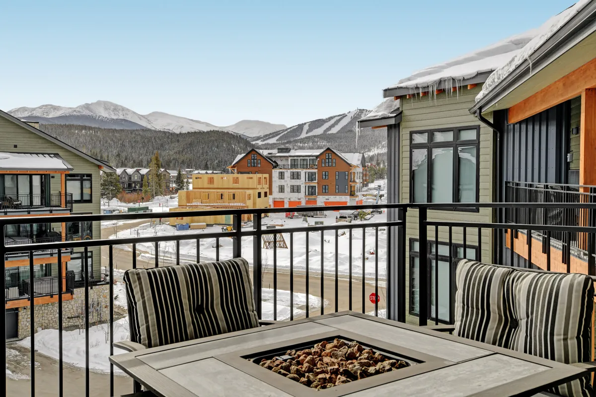 An exterior shot featuring a private balcony with comfortable seating overlooking a majestic mountain range. The cozy setup includes a fire pit, ideal for relaxing while taking in the panoramic views. The scene is set during twilight, highlighting the serene and inviting atmosphere.