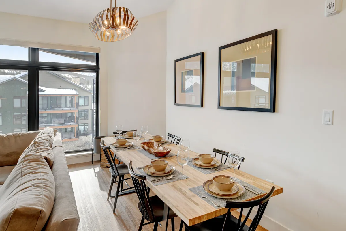 A warm dining area with a six-person table, elegant chandelier, comfortable sofa, and a window framing a picturesque view. The setup is ideal for family feasts, game nights, and relaxed gatherings, reinforcing the central hub concept.