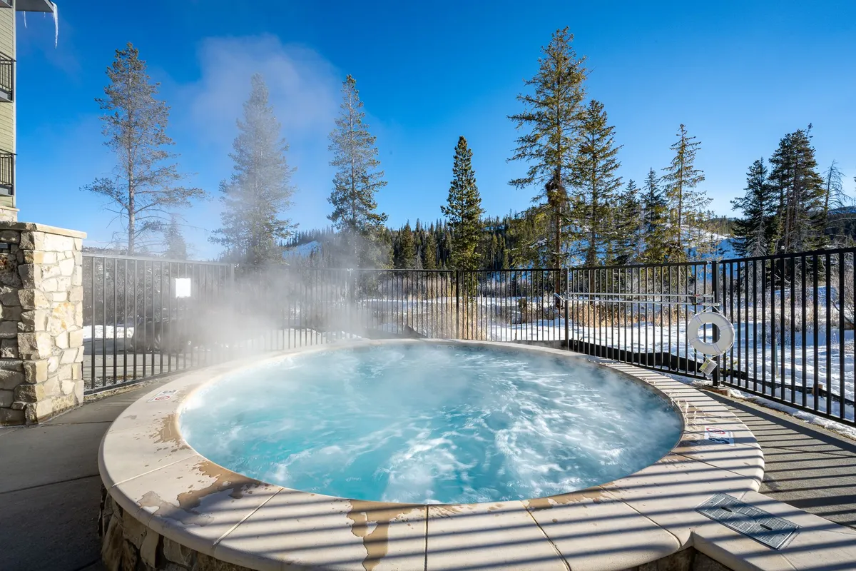 An outdoor hot tub sits amidst pine trees with a panoramic mountain view. Steam rises gently from the water, suggesting relaxation after an active day. The setting is tranquil, inviting, and distinctly tied to the mountain getaway experience.
