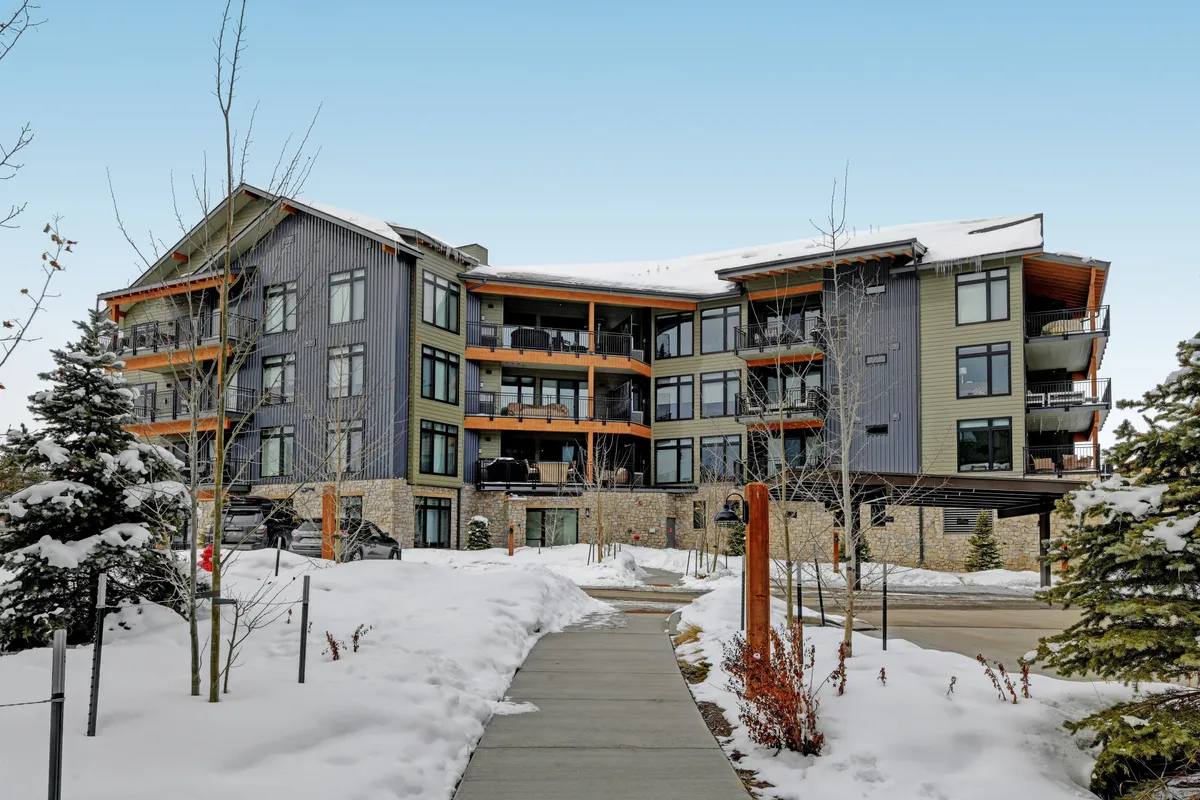 An exterior shot of the condo building, featuring modern architecture with large windows and wooden accents, set against a snowy landscape. The scene suggests warmth and comfort while highlighting proximity to outdoor winter fun.