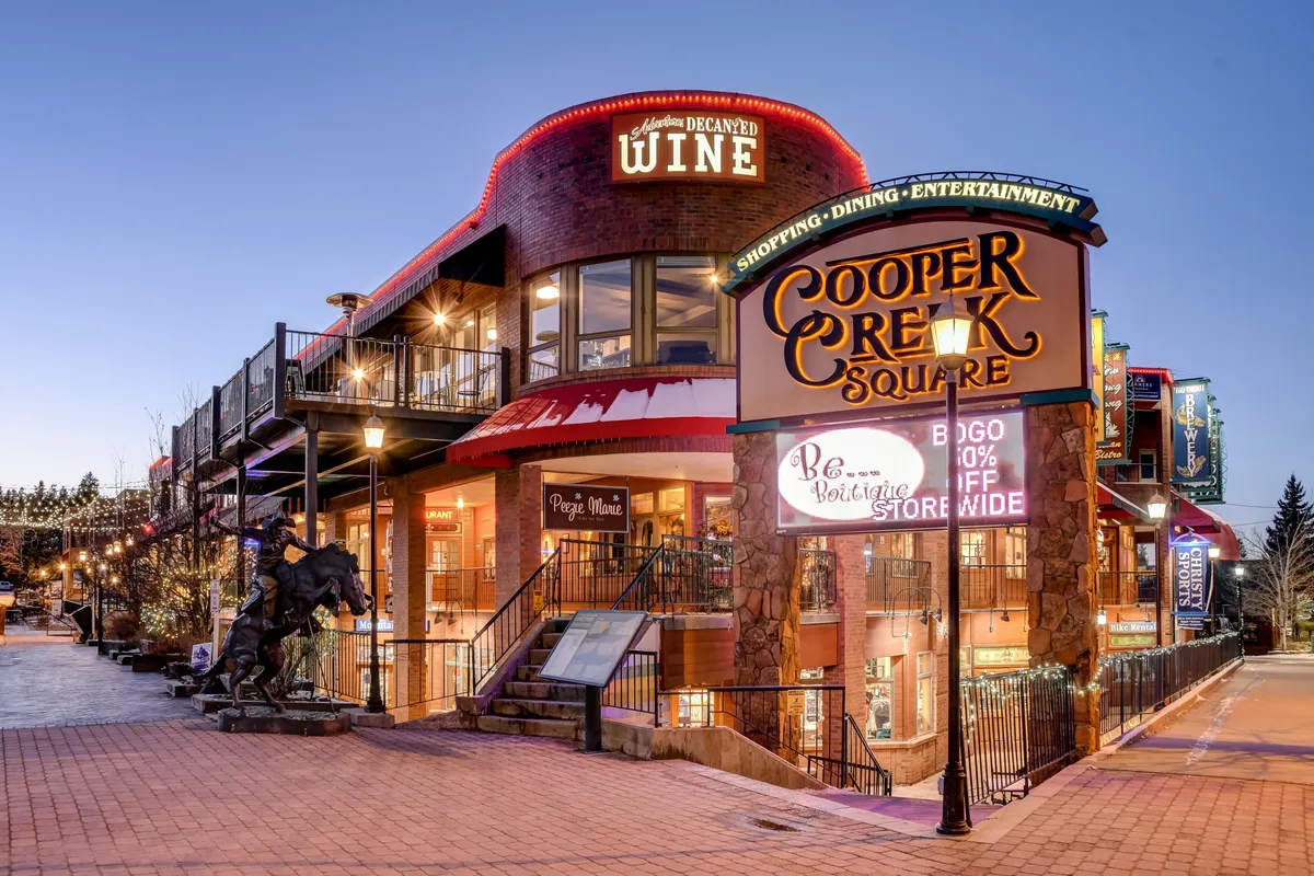 The image showcases the lively Cooper Creek Square with its decorative lighting, a prominent wine shop entrance, a statue, and people milling about. The scene exudes a festive, bustling atmosphere typical of a vibrant downtown area, hinting at diverse dining, shopping, and social opportunities.