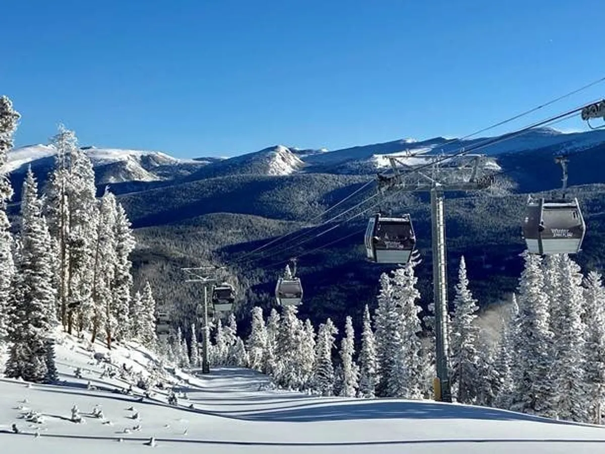 A sweeping exterior view of gondolas gliding over a snow-covered landscape, framed by tall evergreen trees and majestic mountain peaks in the distance. The image radiates the essence of winter adventure and the natural beauty that surrounds Winter Park Resort.