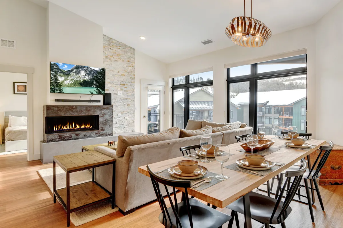 A cozy, stylish living area with a comfortable sofa, elegant dining table set for two, a glowing fireplace, and expansive windows framing beautiful mountain views. The ambiance is warm and intimate, perfect for romantic evenings and relaxation.