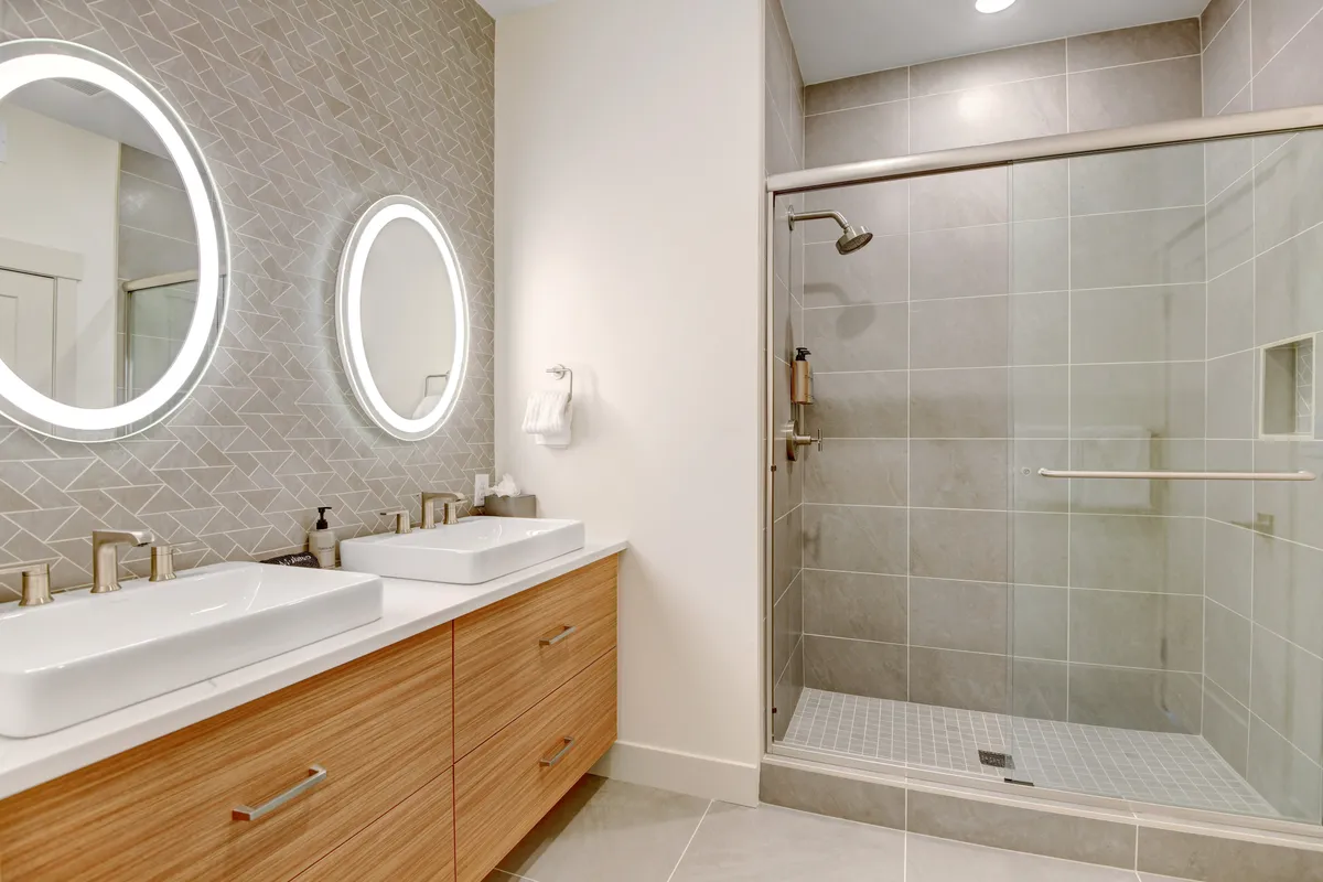 A modern bathroom with double sinks, round illuminated mirrors, and a glass-enclosed shower. The space is bright, clean, and exudes a spa-like atmosphere, echoing the description of the master suite's ensuite.