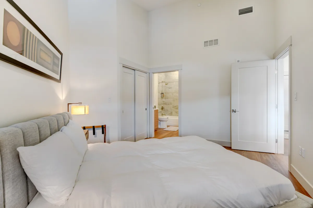 A tranquil master bedroom with a king-sized bed dressed in white premium linens, modern artwork on the wall, and a doorway that leads to an ensuite bathroom. The serene mood and layout emphasize relaxation and comfort, ideal for restful nights and private retreats.