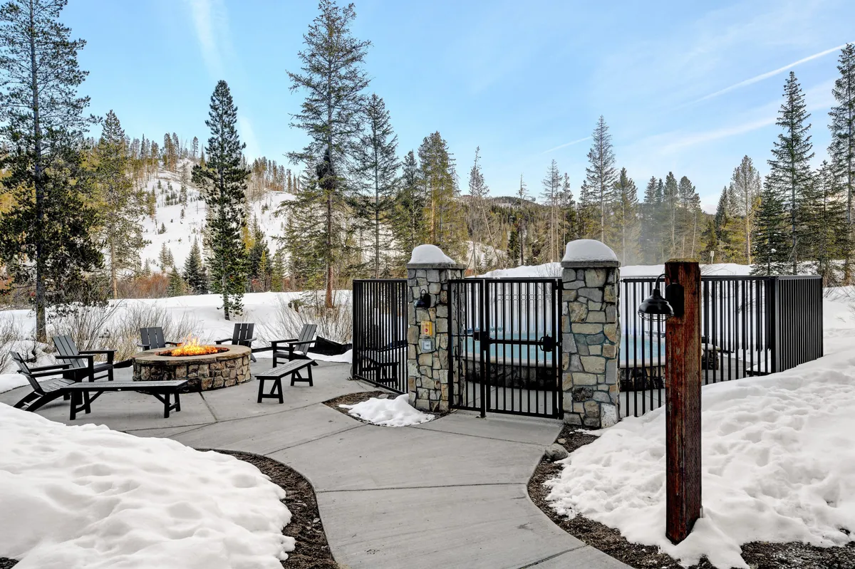 An inviting exterior scene features both a steaming hot tub and a glowing fire pit surrounded by snow and pine trees. The area is arranged for social gatherings and relaxation, with seating encircling the fire and the hot tub offering a tranquil spot to soak. The backdrop of snow and trees underscores the connection to nature and the mountain environment.