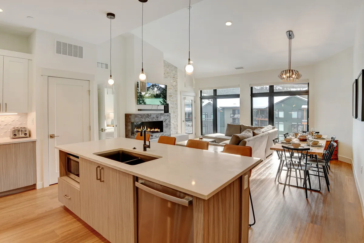 This image features a welcoming open-concept space that seamlessly connects a modern kitchen with a large island, an adjacent dining table with seating for six, a cozy fireplace, and expansive windows that showcase mountain views. The natural light flooding in highlights the contemporary finishes, creating an inviting hub for gatherings.