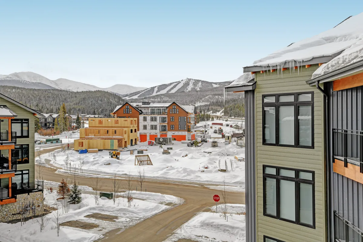 A broad exterior view of snow-covered mountains, newly constructed buildings, and a welcoming community roadway. The scene suggests easy access to mountain sports and community amenities, with an inviting atmosphere.