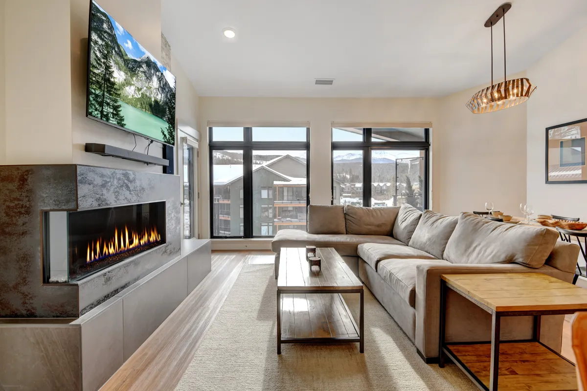 The image features a spacious, light-filled living room with a large sectional sofa arranged around a modern fireplace and a flat-screen TV. Expansive windows offer stunning scenic views, while a wooden coffee table adds a touch of rustic charm. The inviting setup is perfect for groups to relax, socialize, and take in the mountain sunsets after a day of adventures in Winter Park.