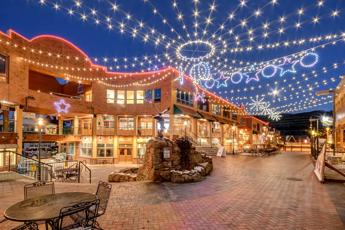 A magical evening scene shows twinkling lights and decorative stars strung above a central fountain and brick pathway, set amid bustling shops and restaurants. People stroll and gather, evoking a sense of community celebration and romance.